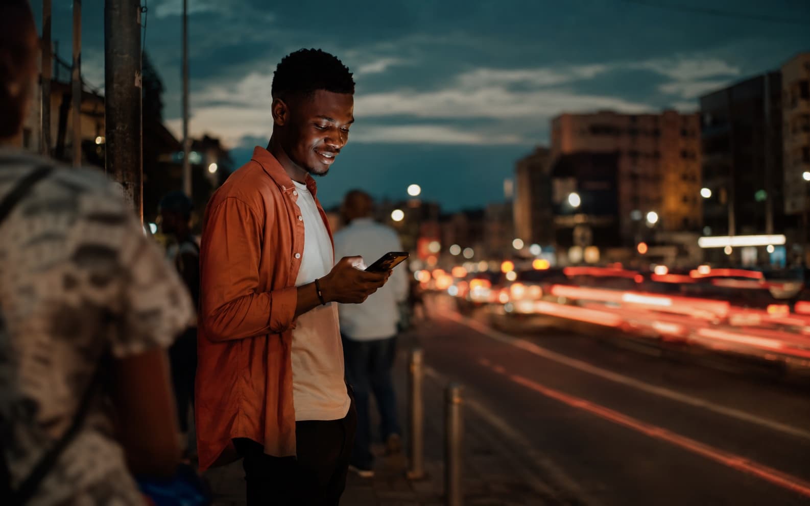 Man on a busy kerb at dusk comparing ride offers on his phone